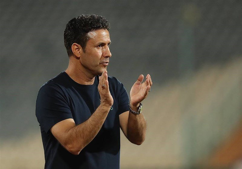 A man in a black t-shirt claps his hands while standing on the sidelines of an outdoor field at night.