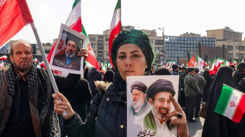 Crowd at an outdoor rally waves Iranian flags and holds portraits of men in clerical attire and religious headwear.