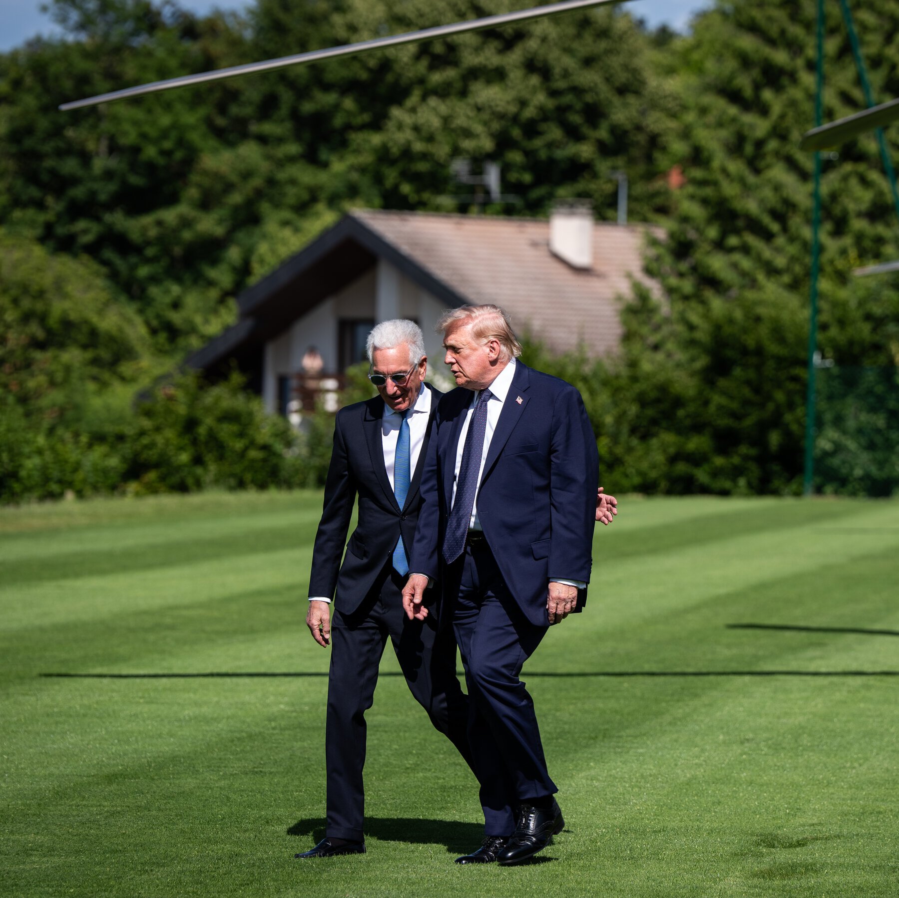 Two men in dark suits walk together across a manicured green lawn, with a house and forested hillside visible in the background.