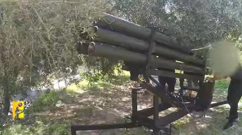A multiple-barrel rocket launcher mounted on a metal frame is positioned among trees in an outdoor setting, with a partially visible figure standing nearby.