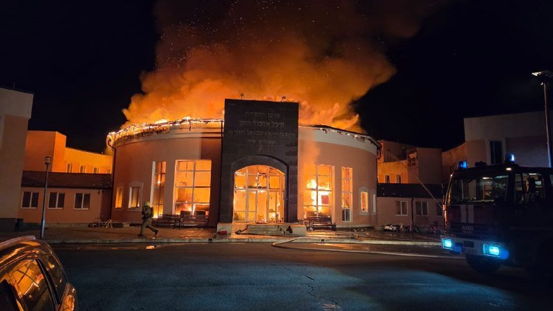 A large circular building is engulfed in flames and thick smoke at night, with a fire truck parked nearby and a person walking across the foreground.