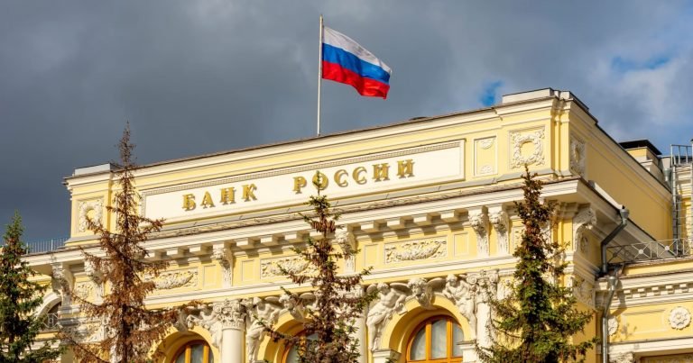 A Russian flag flies atop an ornate yellow building displaying Cyrillic text reading "Bank of Russia."