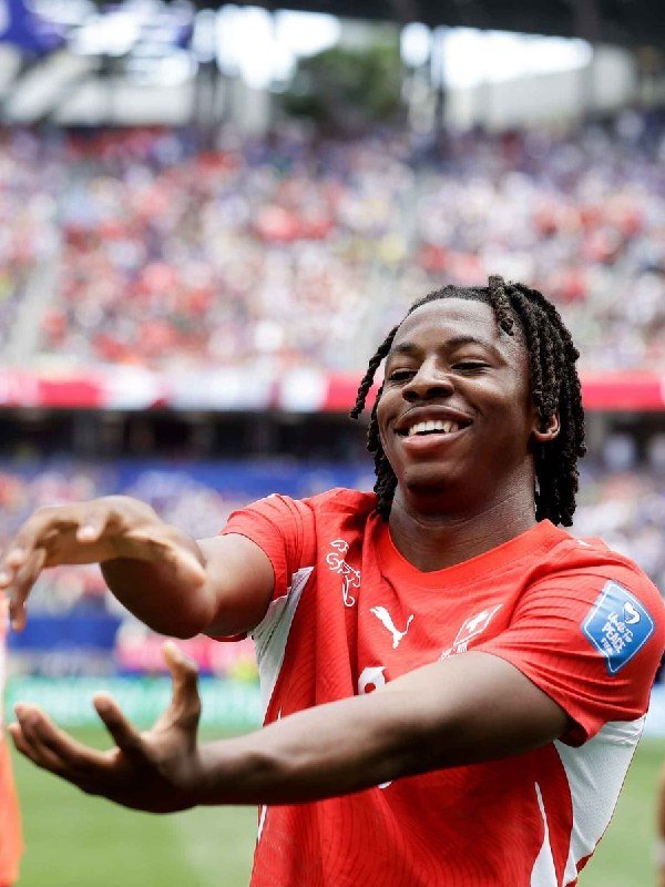 A smiling soccer player with braided hair, wearing a red jersey, celebrates with outstretched arms in a stadium filled with spectators.