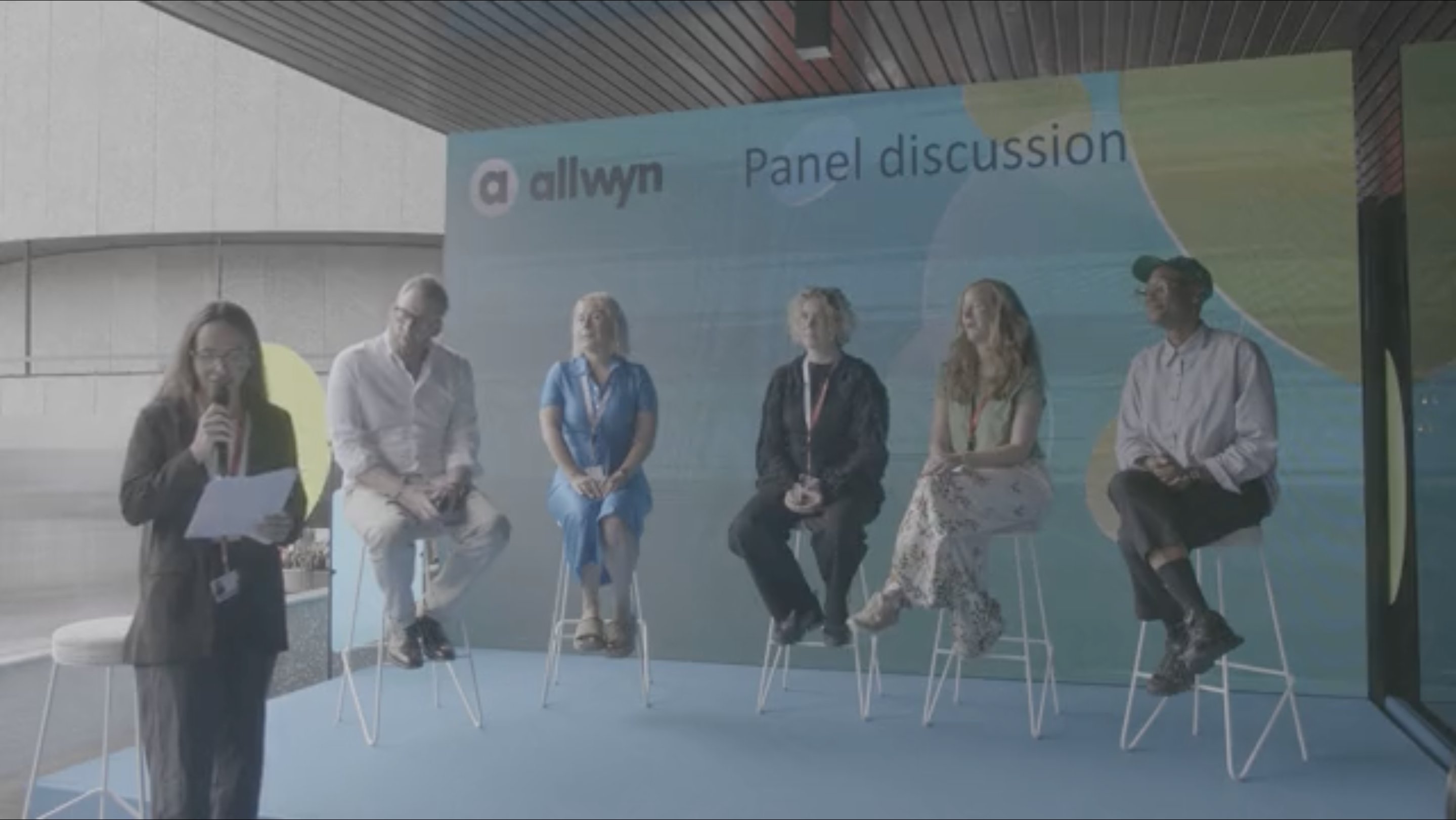 Six people sit on stools on a stage before an "allwyn Panel discussion" backdrop, while a seventh person stands holding a microphone and papers.