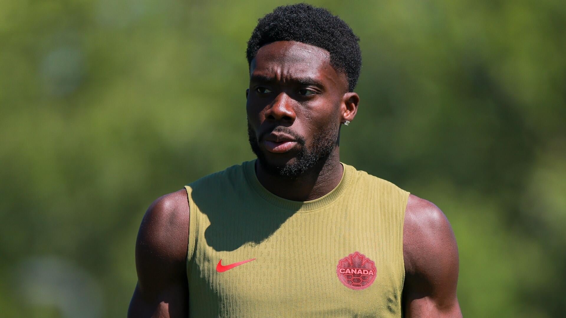 A muscular man wearing an olive sleeveless Nike shirt with a red "Canada" crest stands outdoors against a blurred green background.
