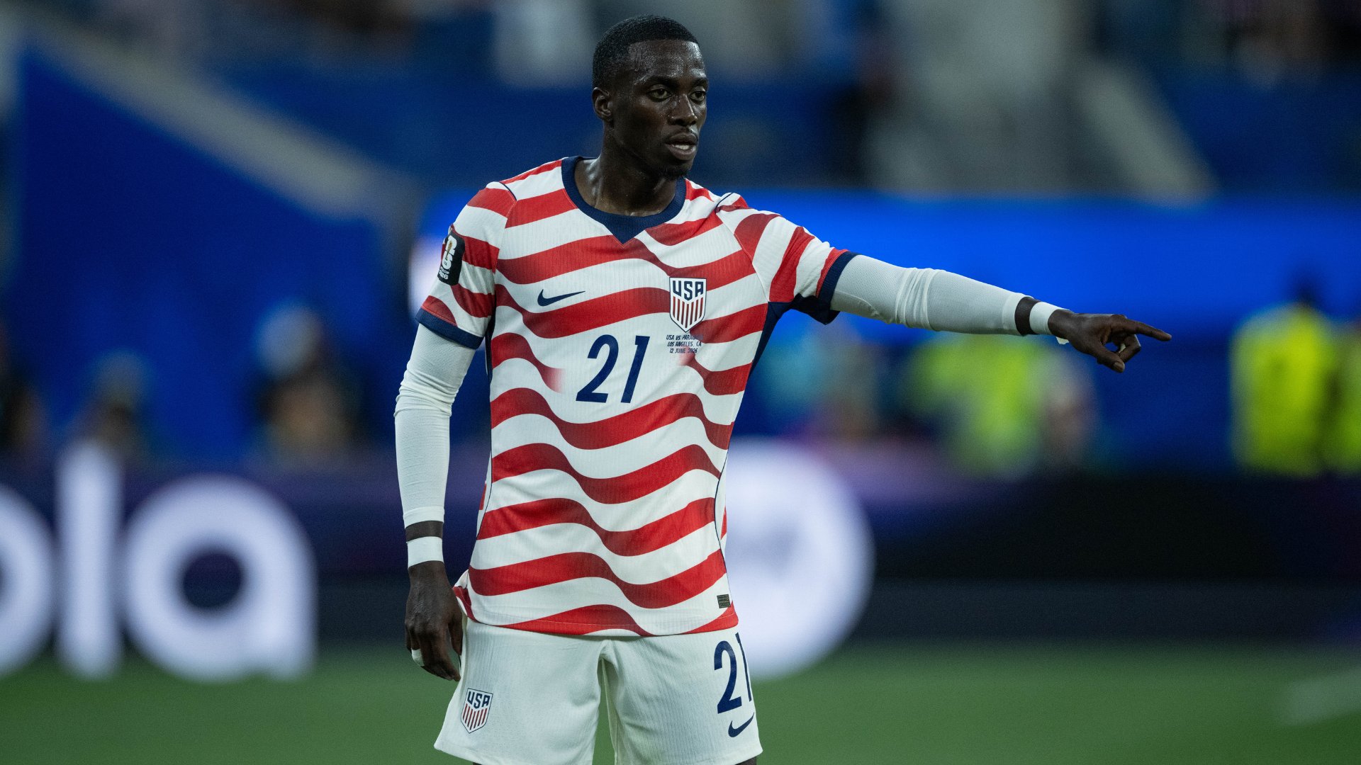A soccer player in a red-and-white striped USMNT jersey, number 21, points to the right during a match on a stadium field.