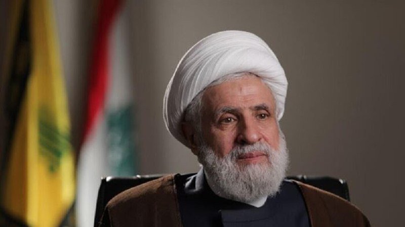 A bearded cleric in a white turban and dark robes sits indoors with yellow and red-white-green flags visible in the background.