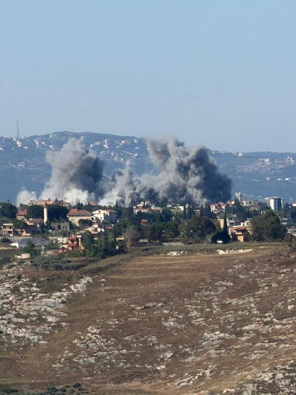 Large plumes of dark smoke rise above a residential area with red-roofed buildings, viewed across a rocky, arid foreground.