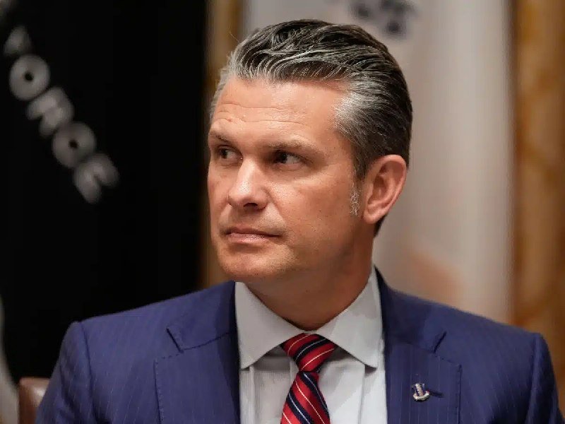 A man in a dark pinstripe suit with a red striped tie and an American flag lapel pin looks off-camera during a formal indoor setting.