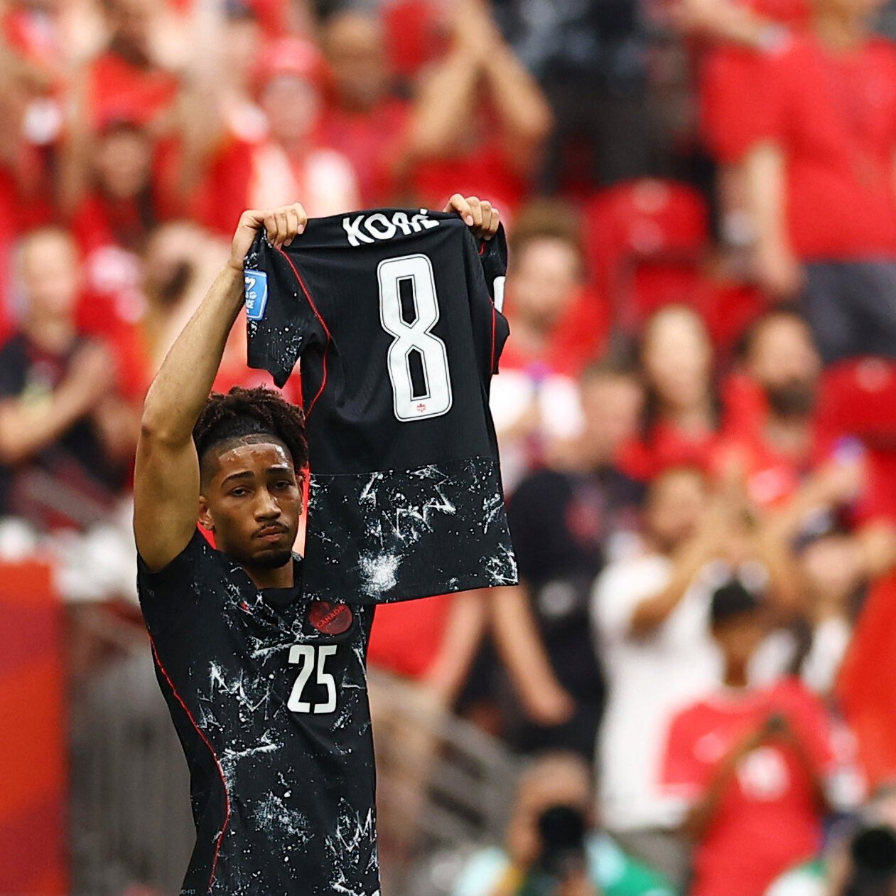 A soccer player wearing a black jersey with the number 25 holds up a black jersey reading "KOBE" and the number 8, facing a crowd in red.