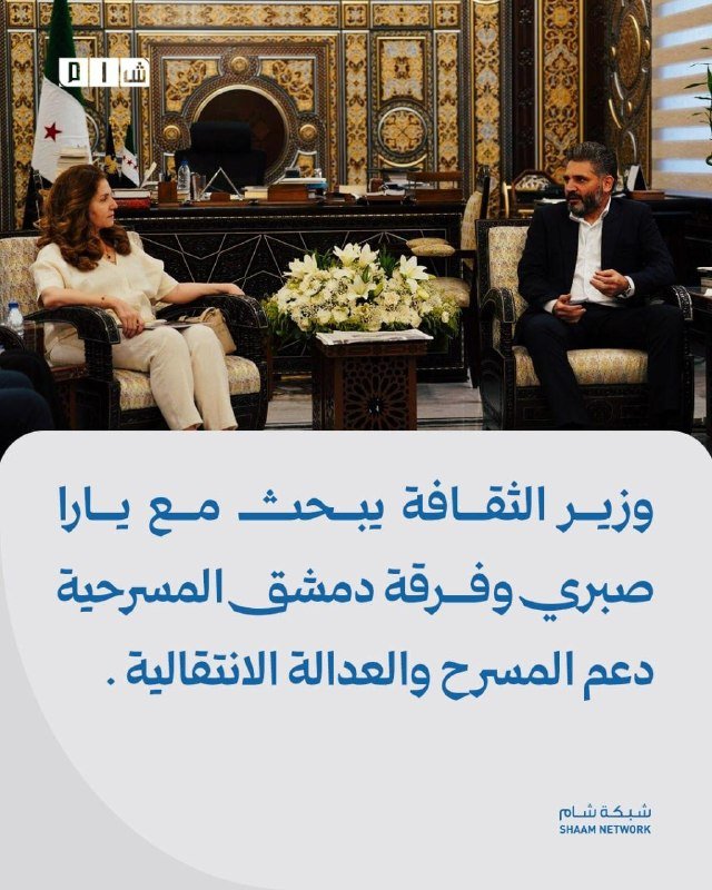 A man and woman sit facing each other in ornate chairs in a decorated room with a Syrian flag, accompanied by Arabic text identifying it as a cultural ministry post.