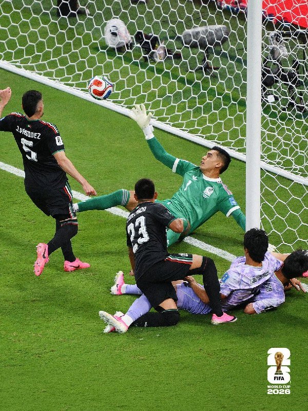 A goalkeeper in green dives to block a shot near the goalpost as opposing players in black and white jerseys challenge during a soccer match.