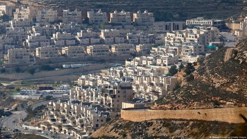 Tan-colored multi-story residential buildings densely cover a hillside in an arid, rocky landscape, with a curving road running along the lower edge.