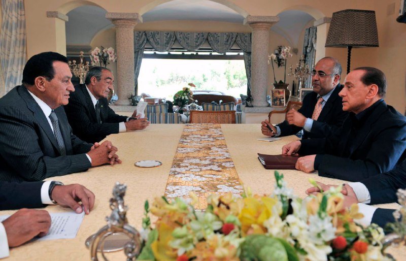 Six men in dark suits sit around a long table adorned with a patterned runner and floral centerpiece in an ornately decorated room with columns and draped windows.