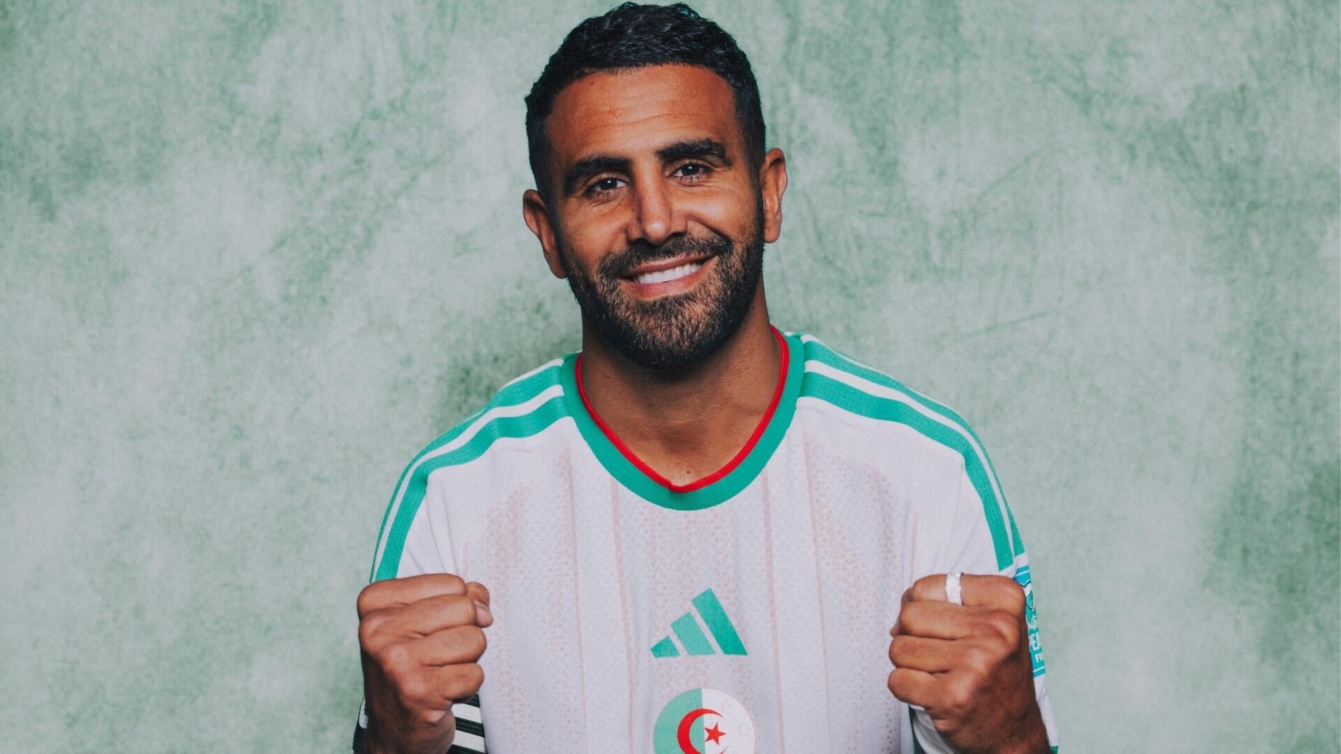 A bearded man smiles and clenches both fists in front of a green textured wall, wearing a white and green soccer jersey featuring the Algerian flag emblem.