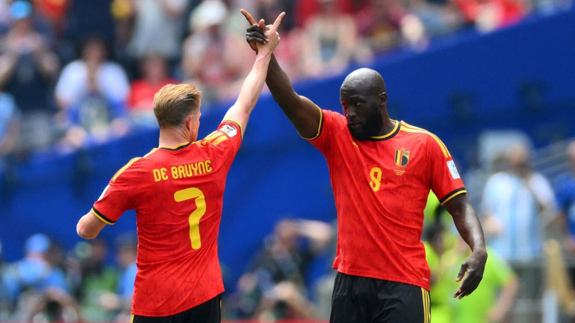Two soccer players in red Belgium jerseys, numbers 7 (De Bruyne) and 9, raise their joined hands in celebration before a stadium crowd.