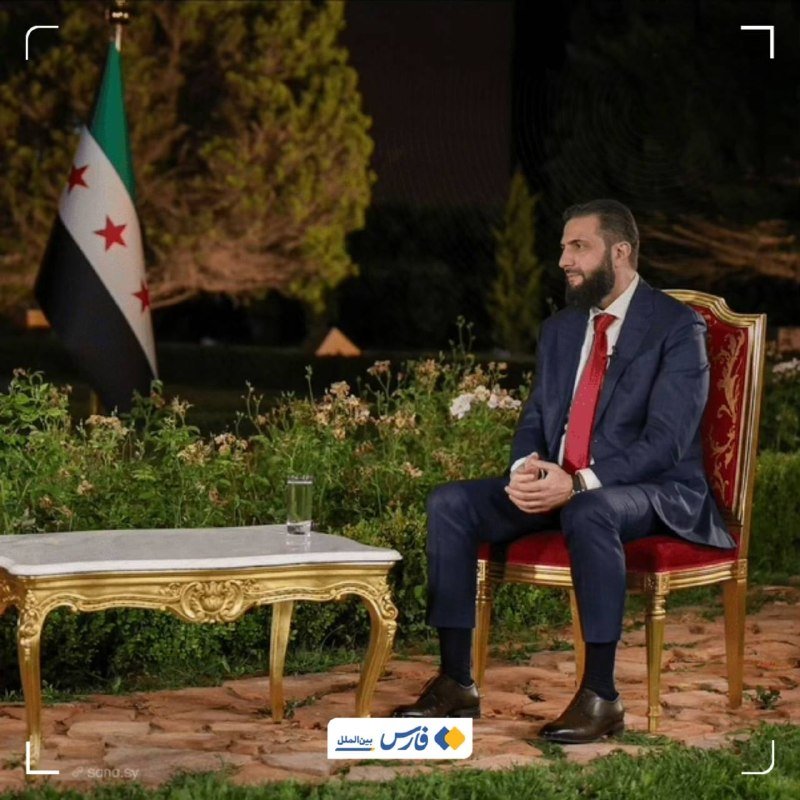 A bearded man in a blue suit and red tie sits on an ornate red chair outdoors at night, beside a Syrian flag and a gilded coffee table.