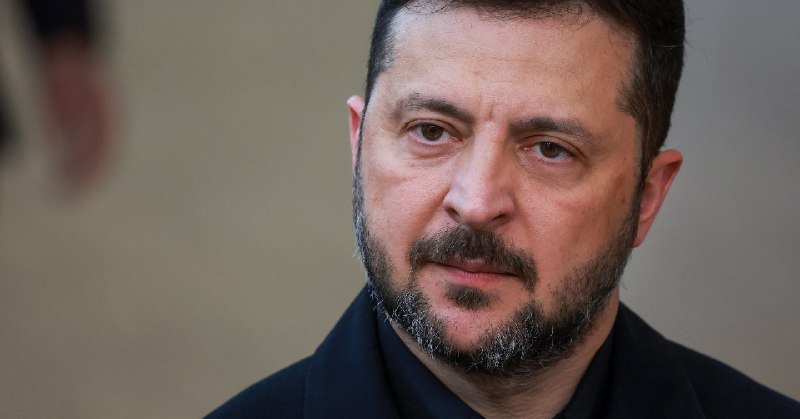 A bearded man in a dark coat looks off to the side with a serious expression against a blurred background.