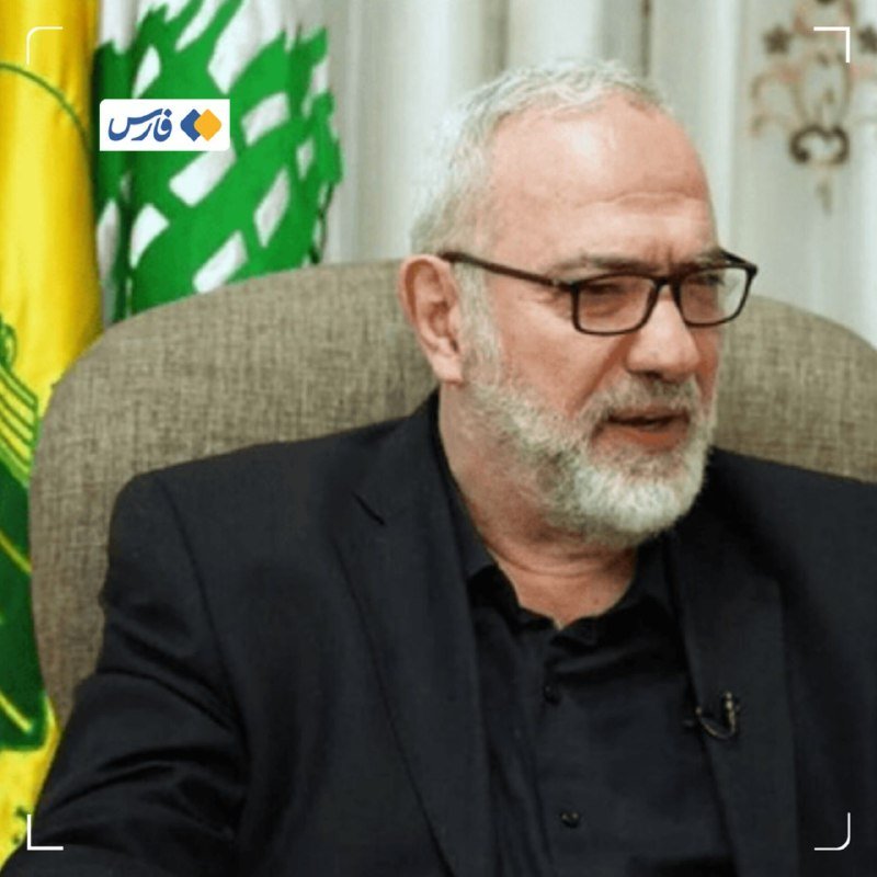 A gray-bearded man wearing glasses and a black shirt sits in a chair beside a green, white, and yellow flag, appearing to speak during an interview.