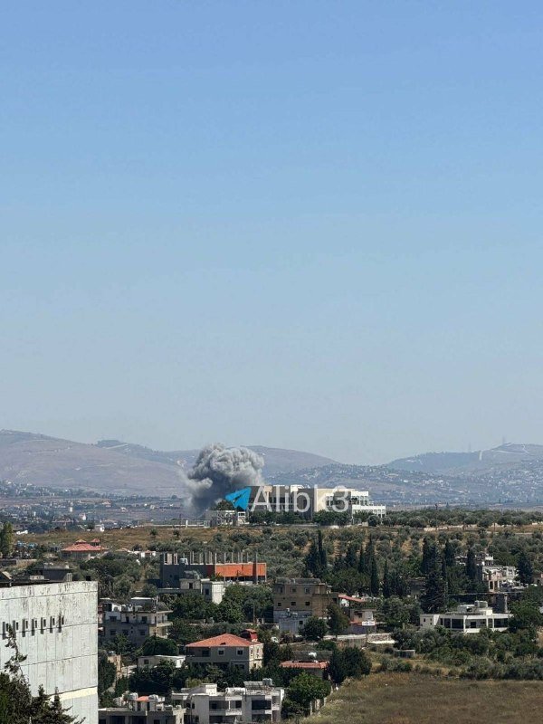 A plume of gray smoke rises above a Middle Eastern town with low-rise buildings, olive groves, and distant hills under a clear blue sky.