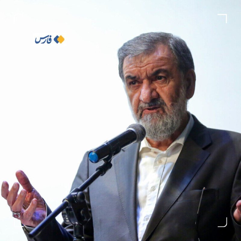 A bearded man in a gray suit speaks into a microphone, gesturing with his hand, against a plain background with a small logo in the corner.