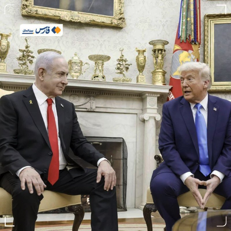 Two men in dark suits sit facing each other in an ornate room with a marble fireplace, gilded decorative urns, and military flags.