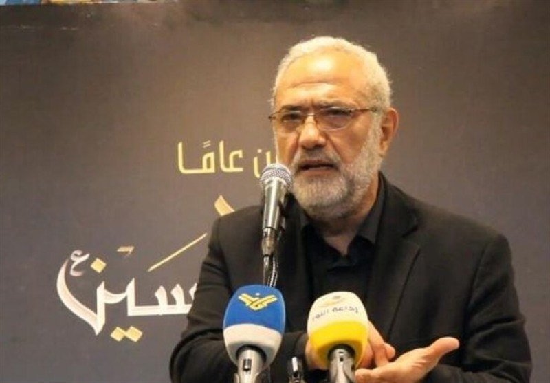 A white-bearded man in a black shirt speaks into multiple microphones against a backdrop featuring Persian script.