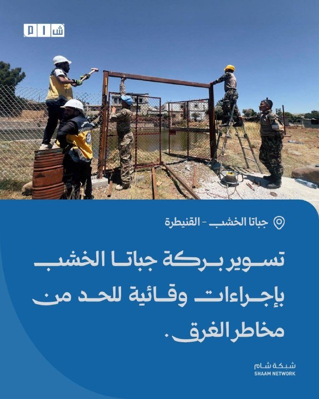 Workers and volunteers repair a metal gate along a fenced area under a clear blue sky, as shown in a Shaam Network news graphic.