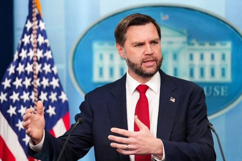A bearded man in a navy suit and red tie gestures while speaking at a podium, with an American flag and a White House seal backdrop visible behind him.