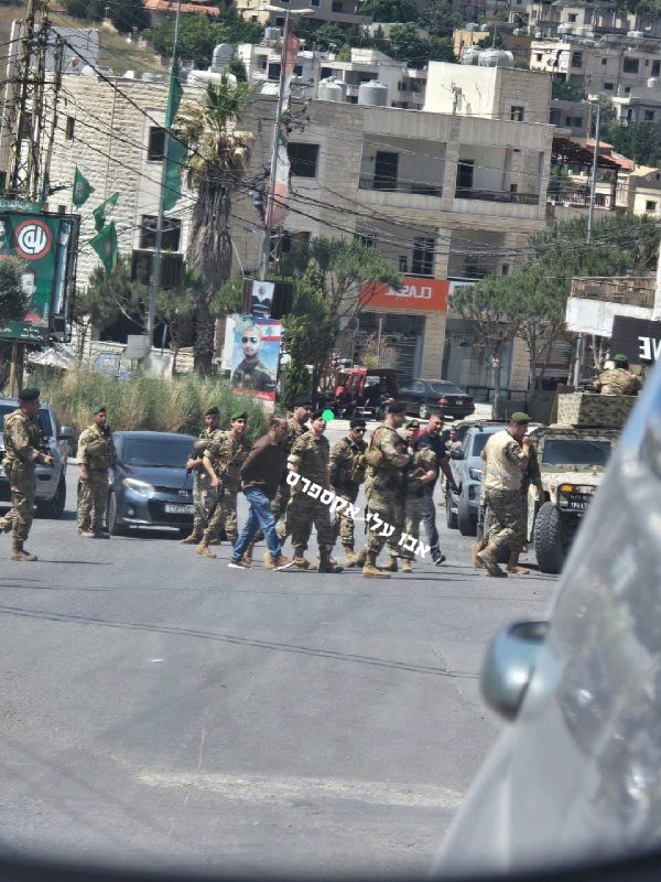Armed soldiers in camouflage uniforms and a civilian walk down a street, escorting a man with covered eyes near a military vehicle, with stone buildings and posters visible in the background.
