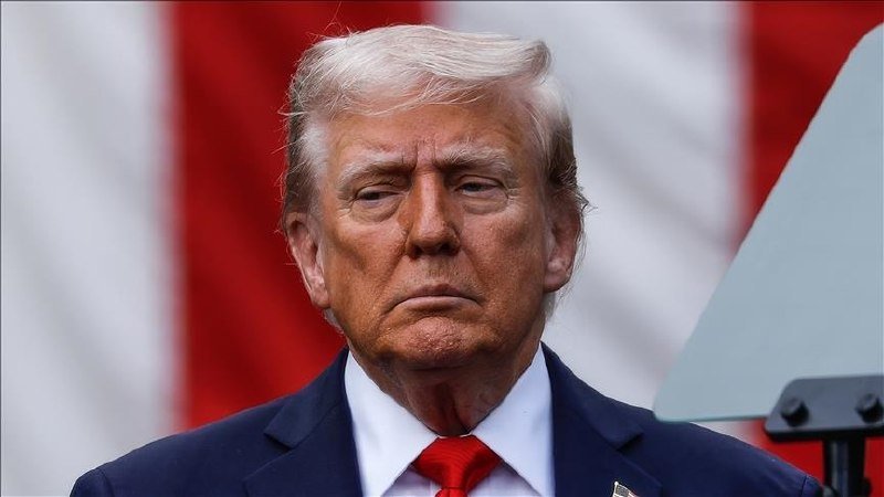 A man with white hair wearing a dark suit, white shirt, and red tie stands in front of an American flag.