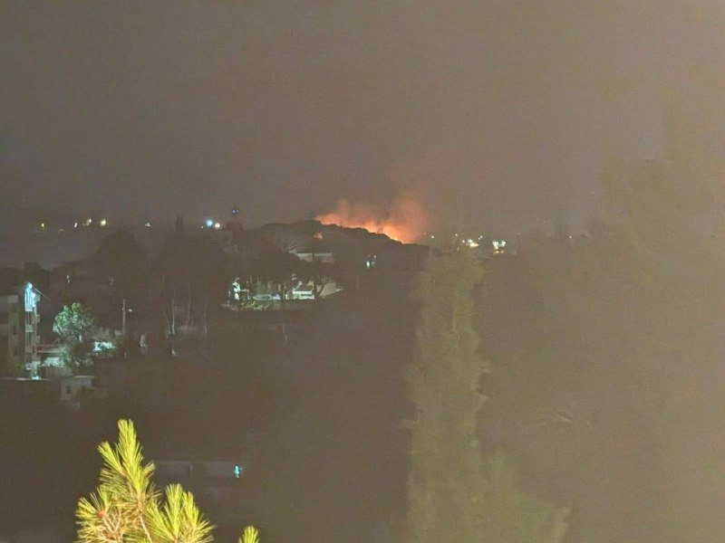 Orange flames and smoke rise above a dark hillside at night, with scattered building lights visible below.