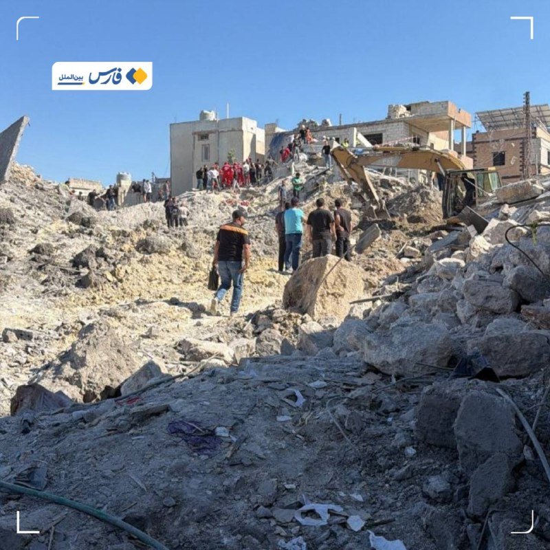 People and a yellow excavator work amid the rubble of destroyed concrete buildings under a clear blue sky, with an Arabic news logo visible in the corner.
