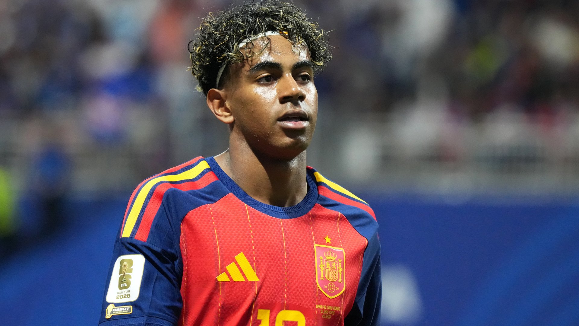 A young soccer player wearing a red Spain national team jersey with a 2026 World Cup qualifier patch stands on the field before a blurred crowd.