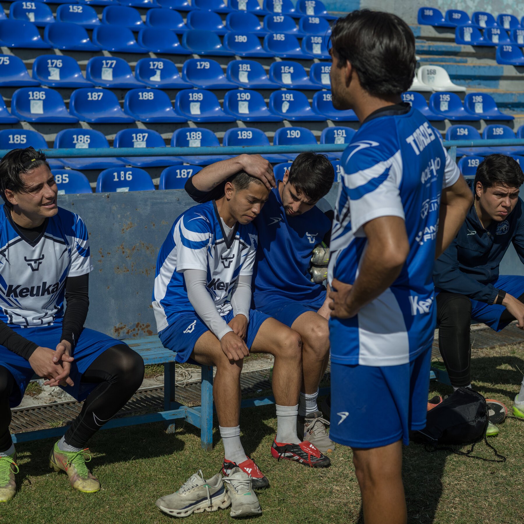 Soccer players in blue and white "Keuka" uniforms sit dejectedly on a bench in front of empty blue stadium seats, while a teammate stands facing them.