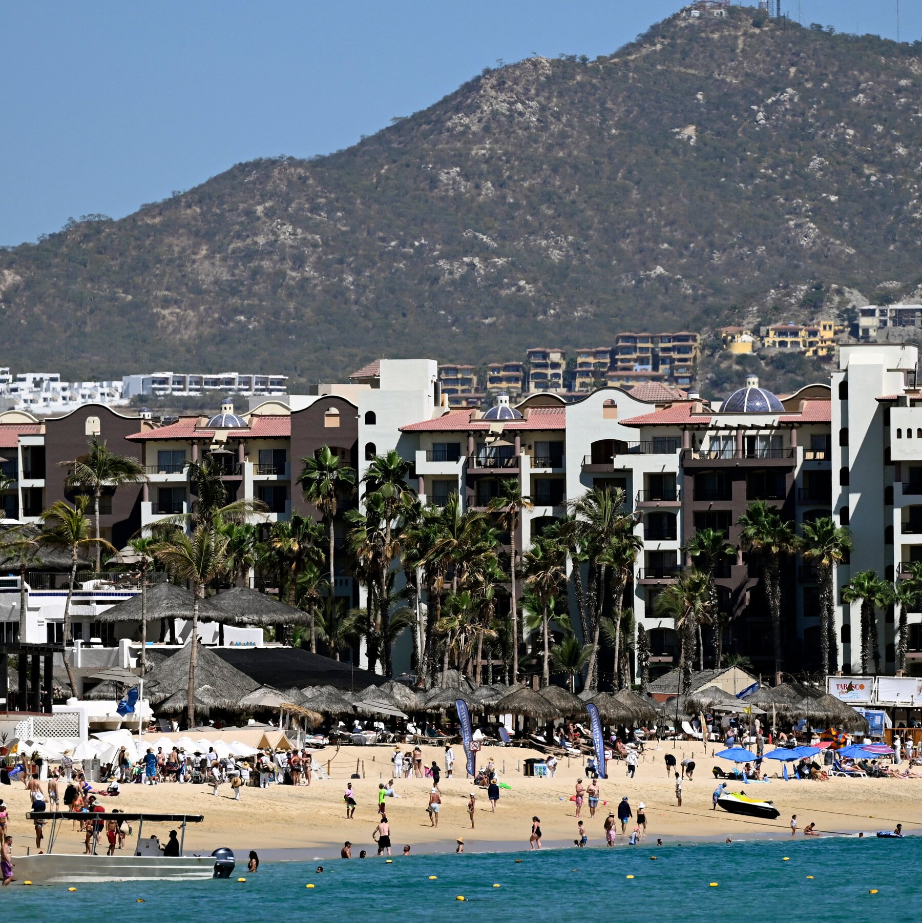Beachgoers gather on a sandy shore beneath palm trees and thatched umbrellas, with multi-story resort buildings and a large rocky mountain rising in the background.