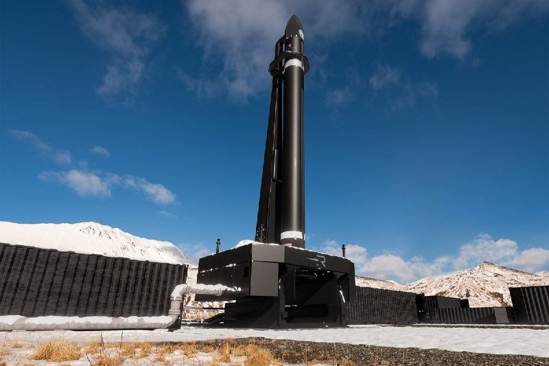 A tall black rocket stands vertically on a launch mount in a snowy, mountainous landscape under a blue sky with scattered clouds.