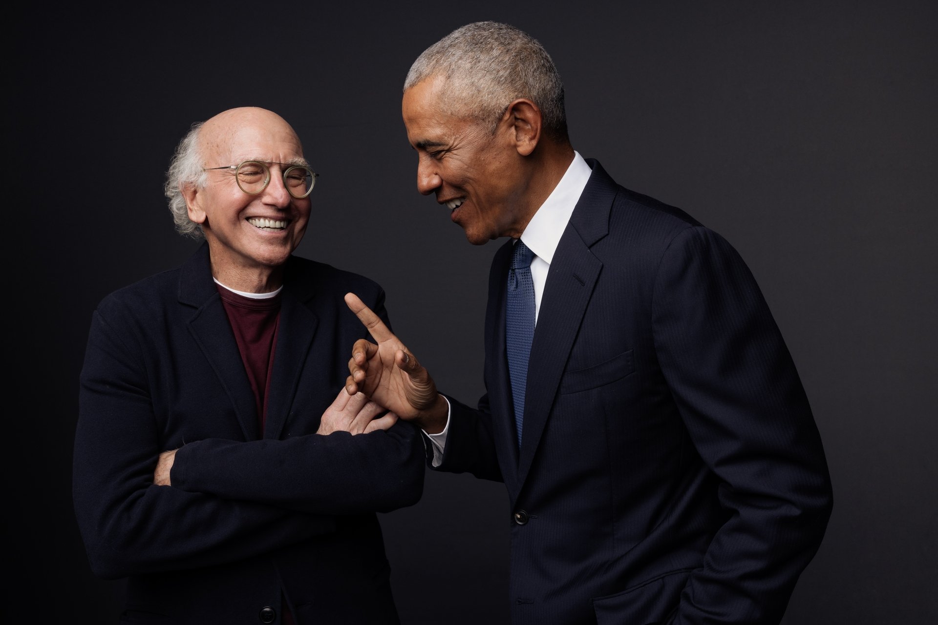 Two men in dark suits smile and laugh together against a black backdrop, one gesturing with his hand toward the other.