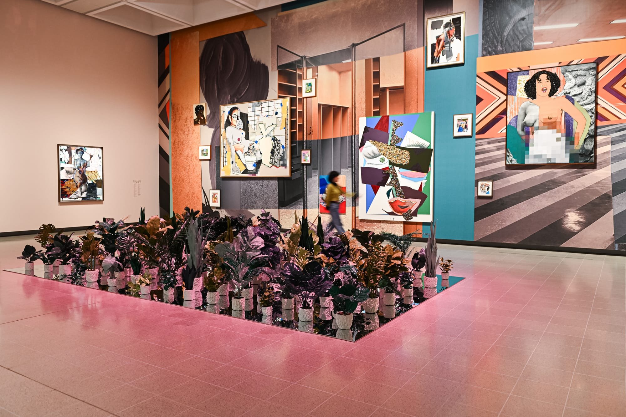 Colorful paintings in mixed-media collage style hang on vibrantly painted gallery walls, displayed above a central mirrored planter filled with potted tropical plants on pink tile flooring.