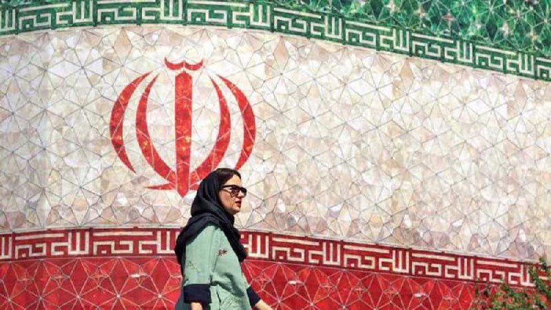 A woman wearing a headscarf and sunglasses walks past a tiled wall displaying a large red national emblem with green and white decorative borders.