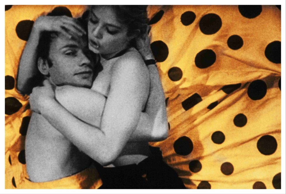 A black-and-white photograph shows two shirtless people embracing against a draped yellow fabric patterned with large black polka dots.