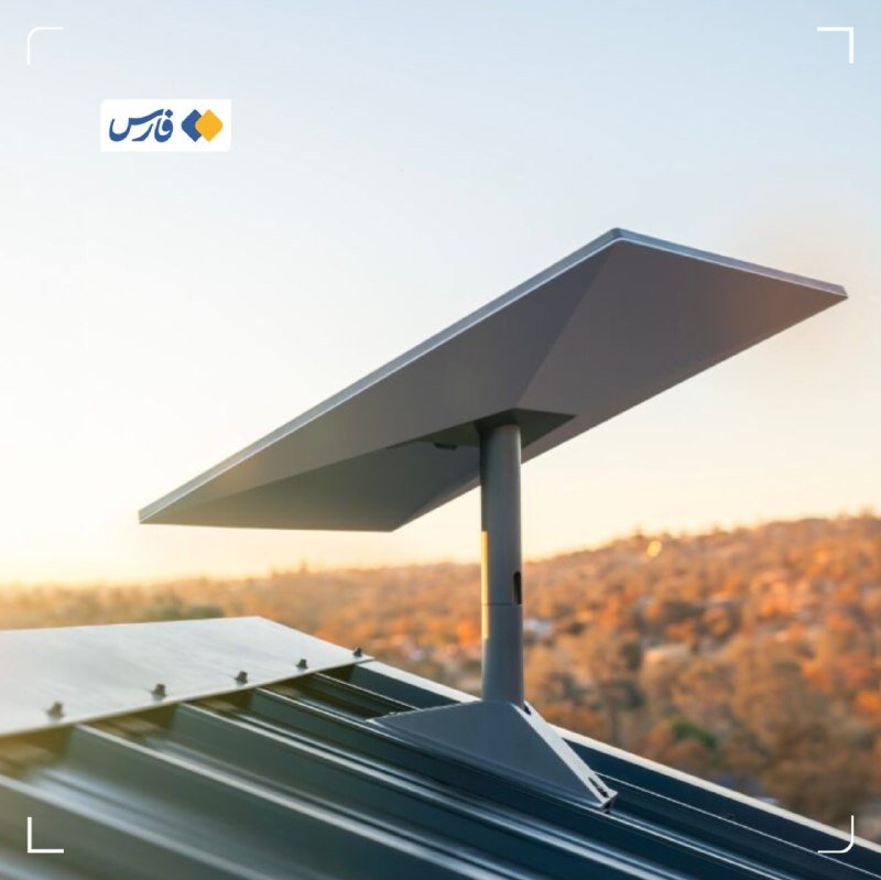 A gray flat satellite dish is mounted on a metal rooftop against a hazy sunset sky with a hilly landscape in the background.