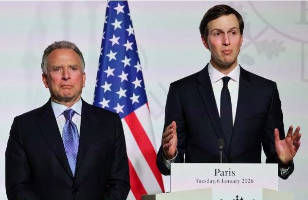 Two men in dark suits stand at a podium labeled "Paris Tuesday, 6 January 2026," with the younger one gesturing while speaking and an American flag behind them.
