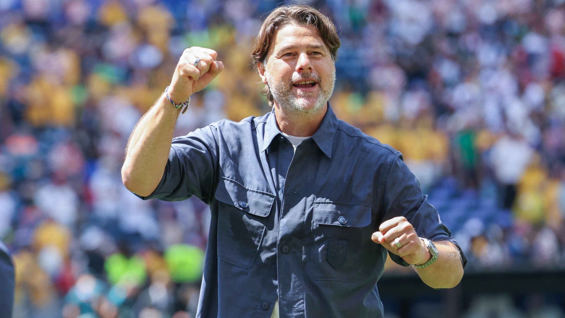 A bearded man in a blue USA soccer shirt raises a clenched fist and smiles while celebrating before a stadium crowd.