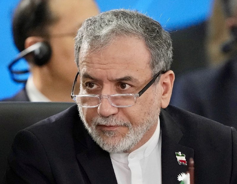 A gray-haired, bearded man wearing glasses and a dark suit with an Iranian flag pin looks forward, with a blurred figure wearing headphones visible behind him.