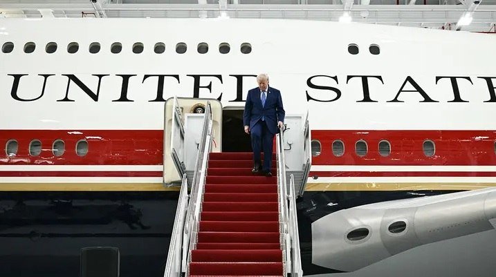 An older man in a dark suit descends a red-carpeted staircase from an aircraft labeled "UNITED STATES" inside a hangar.