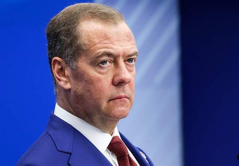 A serious-looking man in a blue suit and patterned tie stands against a blue background with diagonal light streaks.