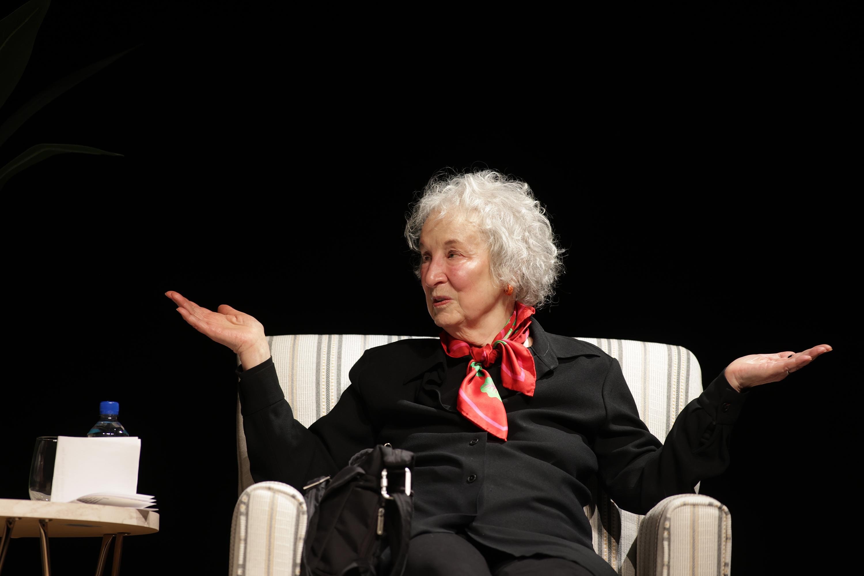 An elderly woman with curly white hair sits in a striped armchair against a dark background, wearing a black shirt and red patterned scarf, with both hands raised palms-up in a gesturing motion.