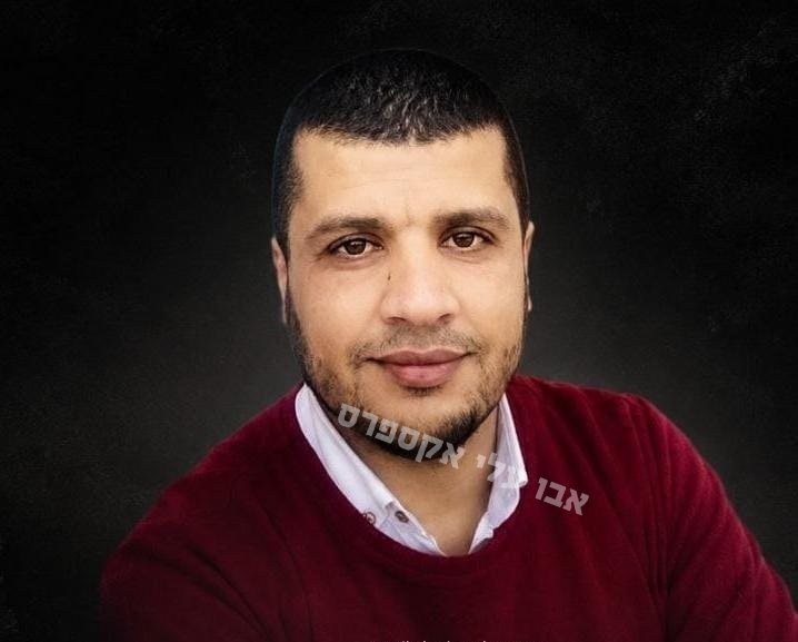 A man with short dark hair and a light beard, wearing a red sweater over a white collared shirt, poses for a portrait against a dark background with Hebrew text overlaid.