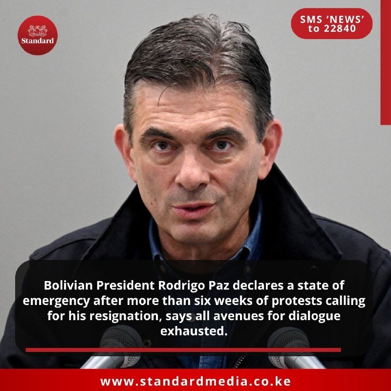 A man in a dark coat speaks at a podium with microphones, with a news graphic overlay featuring "Standard" logo and website URL.